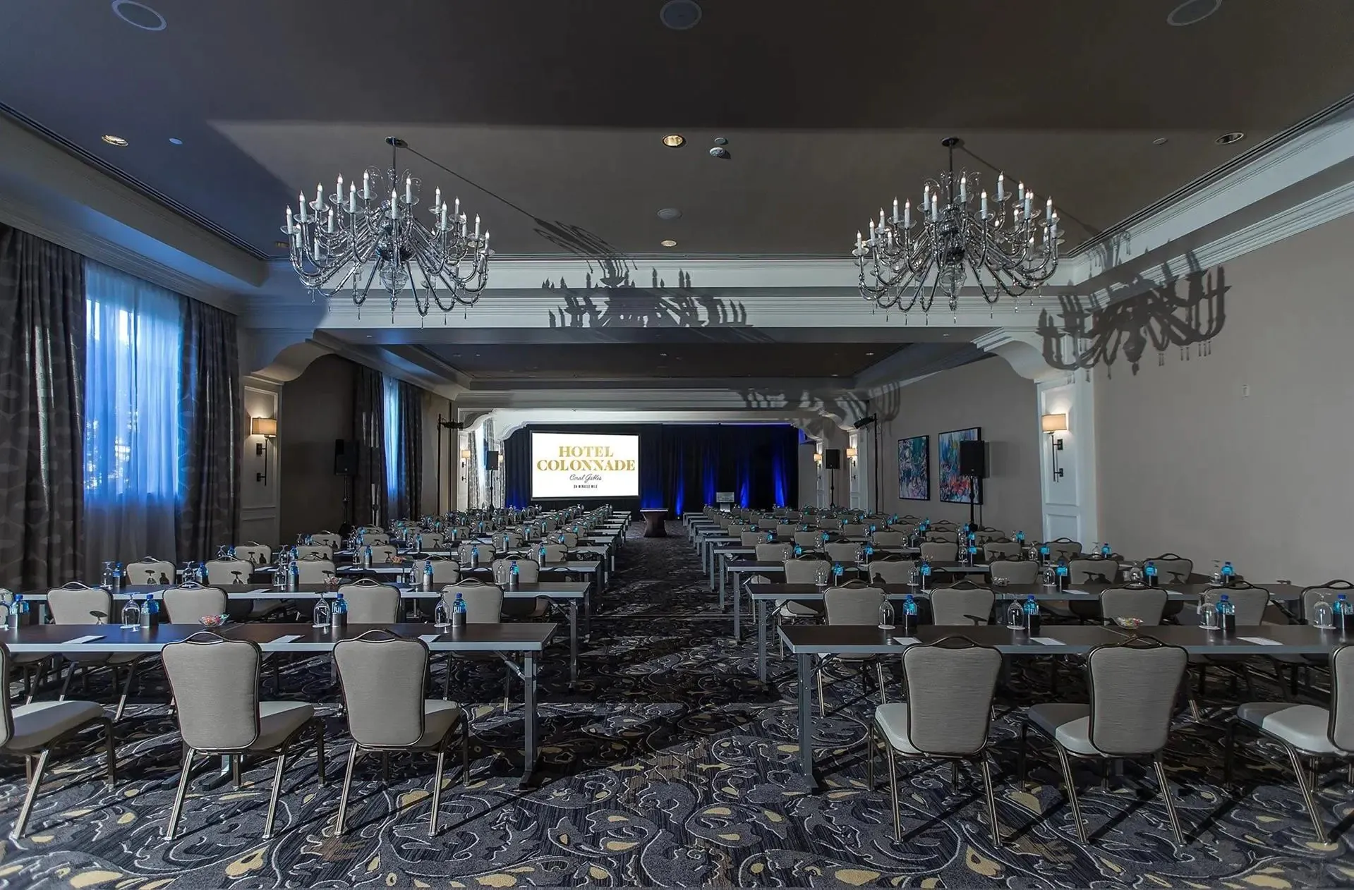 Elegant conference room at Hotel Colonnade with rows of tables and chairs, chandeliers overhead, water bottles on tables, and a large screen at the front.