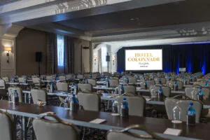 Conference room at Hotel Colonnade with rows of tables and chairs, water bottles, and a large screen at the front displaying HOTEL COLONNADE Coral Gables.