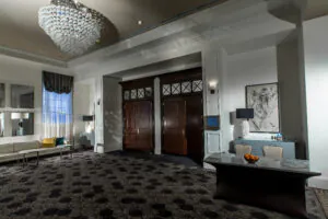 The elegant lobby at Hotel Colonnade features a patterned carpet, marble table with fruit, wall art, wooden double doors, and a modern chandelier overhead.